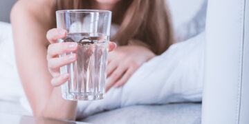 The image depicts a person holding a glass of water, focusing on the hand and the glass. The individual appears to be resting on a bed or couch, with a soft and relaxed ambiance in the background. The glass of water is clear, reflecting light softly.